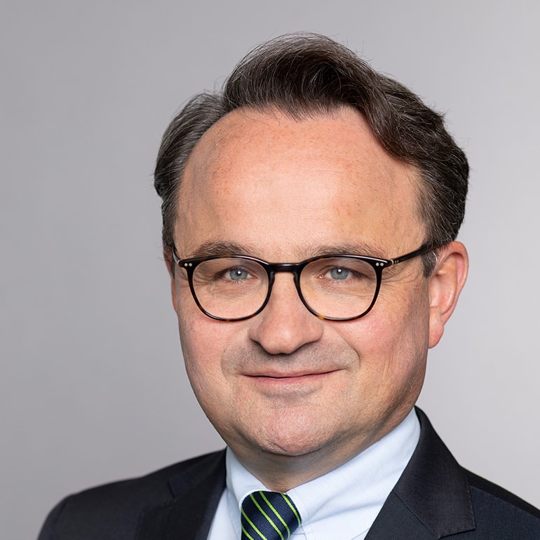 Porträt eines lächelnden Mannes mit Brille, dunklen Haaren und mittellanger Frisur. Er trägt einen Anzug mit einer hellen Hemd und einer gestreiften Krawatte. Der Hintergrund ist neutral grau.