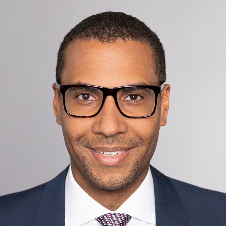 Porträt eines Mannes mit kurzer, dunkler Frisur und Brille. Er trägt einen dunklen Anzug und ein kariertes Hemd. Der Mann lächelt freundlich in die Kamera, vor einem hellen, neutralen Hintergrund.