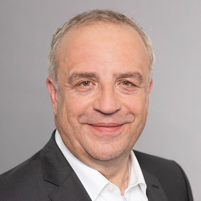 Porträt eines lächelnden Mannes mittleren Alters mit kurzen, grauen Haaren. Er trägt einen dunklen Anzug und ein weißes Hemd. Der Hintergrund ist grau und neutral, was den Fokus auf sein freundliches Gesicht legt.