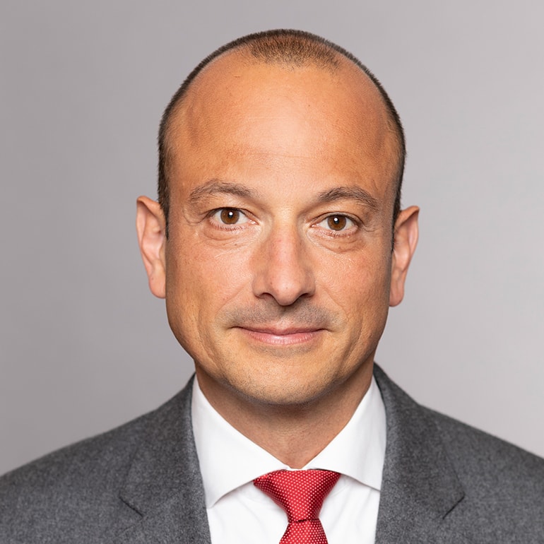 Porträt eines Mannes mit kurzem, glattem Haar, der einen grauen Anzug und eine rote Krawatte trägt. Er hat einen ernsthaften, aber freundlichen Ausdruck und sieht direkt in die Kamera. Der Hintergrund ist einfarbig und neutral.