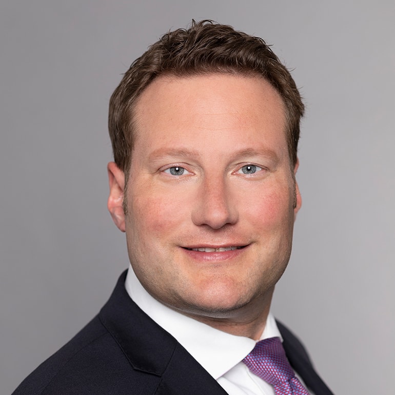 Ein Mann mit kurzen, braunen Haaren lächelt freundlich in die Kamera. Er trägt einen schwarzen Anzug mit einem weißen Hemd und einer lila Krawatte. Der Hintergrund ist neutral und hellgrau, was den Fokus auf sein Gesicht lenkt.