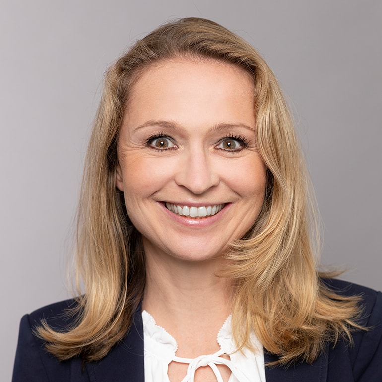 Ein Frau mit schulterlangen, blonden Haaren und einem freundlichen Lächeln. Sie trägt ein dunkelblaues Oberteil und eine weiße Bluse. Der Hintergrund ist grau.