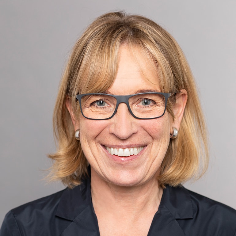 Eine lächelnde Frau mit blonden, schulterlangen Haaren trägt eine schwarze Bluse und braune Brille. Ihr Gesicht strahlt Freundlichkeit aus. Der Hintergrund ist einfarbig grau.