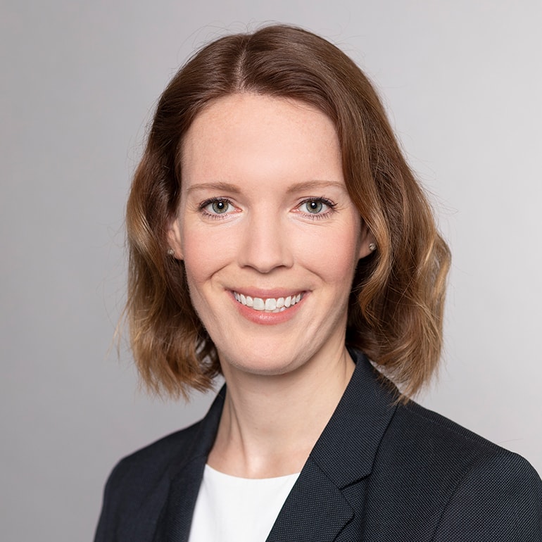 Ein Porträt einer Frau mit schulterlangen, lockigen Haaren und einem freundlichen Lächeln. Sie trägt einen dunklen Blazer über einem weißen Oberteil. Der Hintergrund ist hell und neutral.