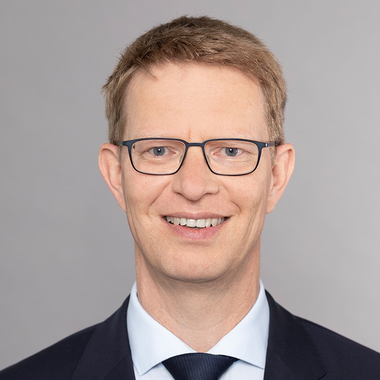 Das Bild zeigt einen Mann mit kurzem, blondem Haar, der in einem Anzug mit Krawatte und weißem Hemd lächelt. Er trägt eine Brille und steht vor einem grauen Hintergrund.