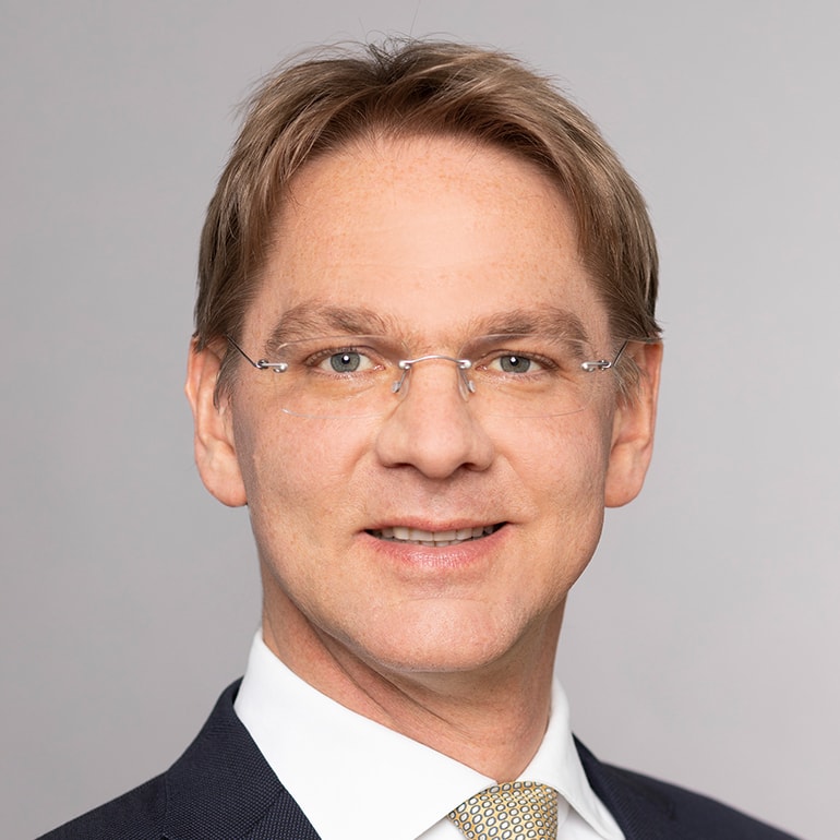 Porträt eines Mannes mit kurzen, blonden Haaren. Er trägt eine Brille und einen Anzug mit einem weißen Hemd und einer beige Krawatte. Der Hintergrund ist einfarbig. Der Mann lächelt freundlich in die Kamera.