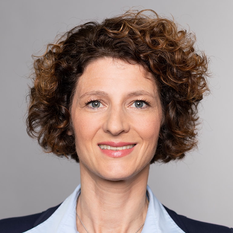 Porträt einer Frau mit lockigem, braunem Haar und strahlendem Lächeln. Sie trägt eine hellblaue Bluse und hat einen freundlichen Blick. Der Hintergrund ist neutral und unauffällig.