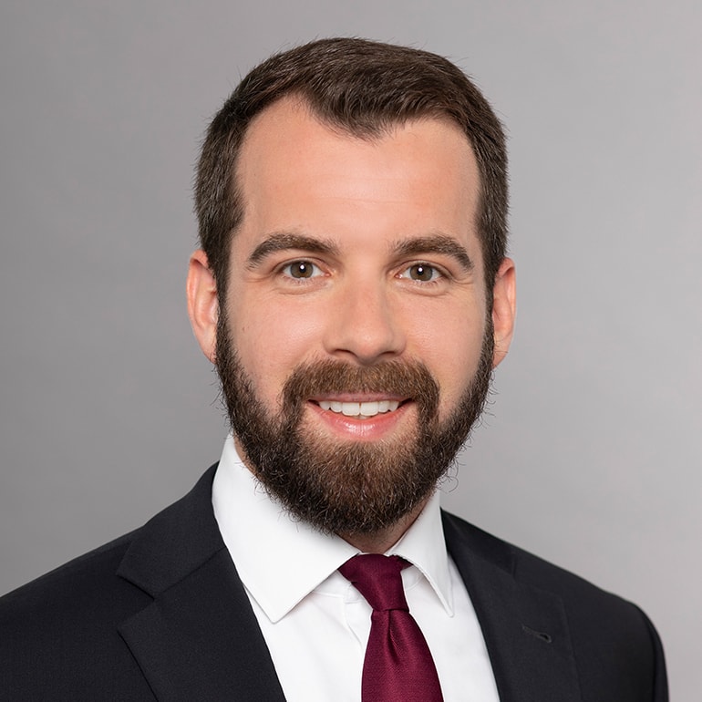 Der Mann trägt einen dunklen Anzug und eine bordeauxfarbene Krawatte. Sein lächelndes Gesicht zeigt einen gepflegten Bart und kurzes, dunkles Haar. Der Hintergrund ist hell und einfarbig, was den Fokus auf seine freundliche Ausstrahlung legt.