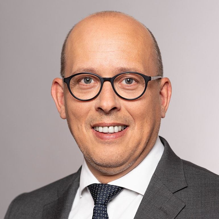 Ein lächelnder Mann mit glattem, kurzer Haar, der eine Brille trägt. Er ist in einem grauen Anzug und einer weißen Hemd mit einer gemusterten Krawatte gekleidet. Der Hintergrund ist hell und neutral.