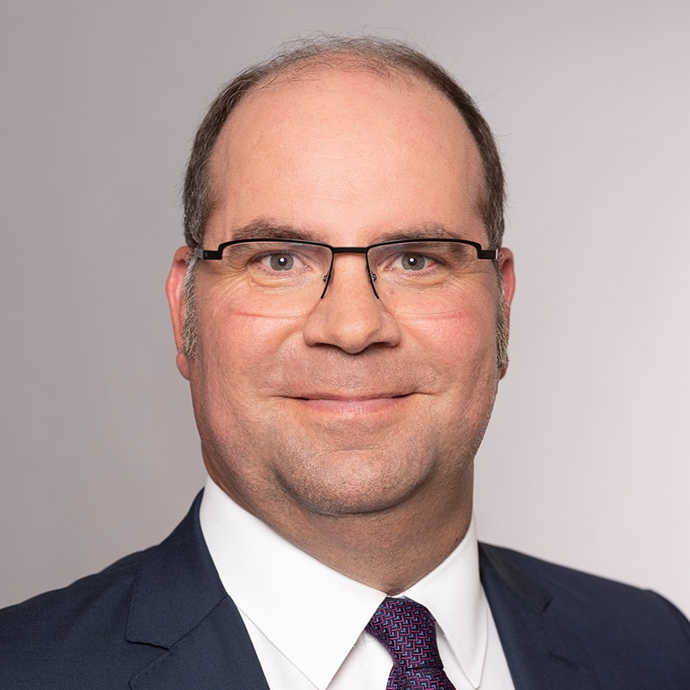 Ein Mann mit Brille und einem freundlichen Lächeln steht vor einem einfarbigen Hintergrund. Er trägt einen dunklen Anzug mit einer hellen Krawatte. Sein Haar ist kurz und hat eine leichte Grau-Note. Die Aufnahme zeigt sein Gesicht bis zur Oberkante der Schultern.