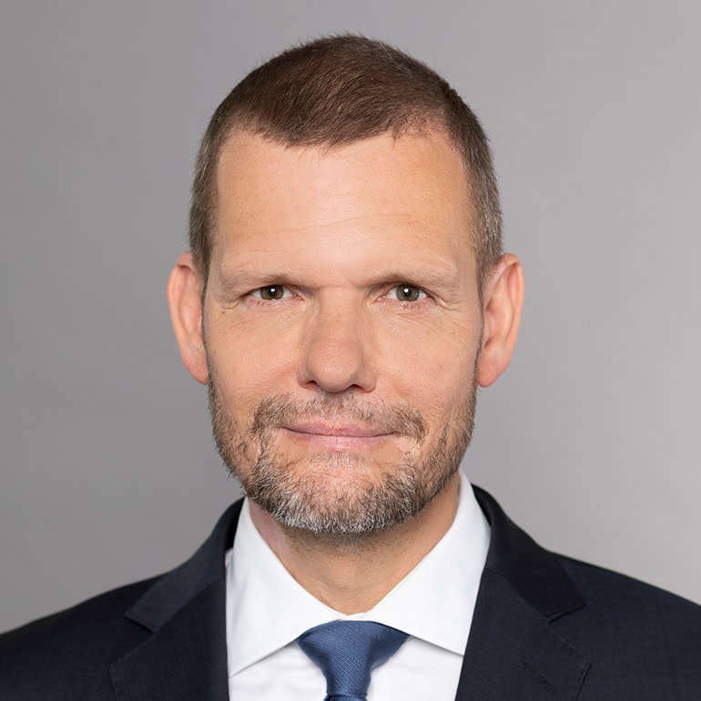 Porträt eines Mannes mit kurzem, graubraunem Haar, einem gepflegten Bart und einem freundlichen Gesichtsausdruck. Er trägt einen dunklen Anzug mit einer weißen Hemd und einer blauen Krawatte. Der Hintergrund ist hell und neutral gehalten.