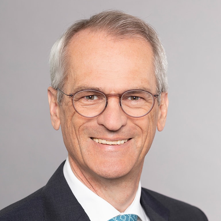 Porträt eines lächelnden Mannes mit grauen Haaren und Brille, der einen eleganten Anzug trägt. Der Hintergrund ist einfarbig hellgrau, was den Fokus auf sein freundliches Gesicht lenkt.