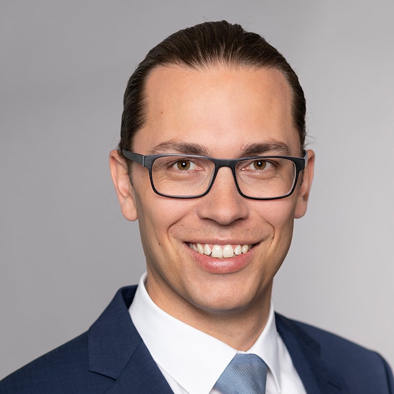 Porträt eines lächelnden Mannes mit Brille, dunkelblonden Haaren, die zurückgebunden sind. Er trägt einen Anzug mit einem weißen Hemd und einer hellblauen Krawatte. Der Hintergrund ist neutral und hell.