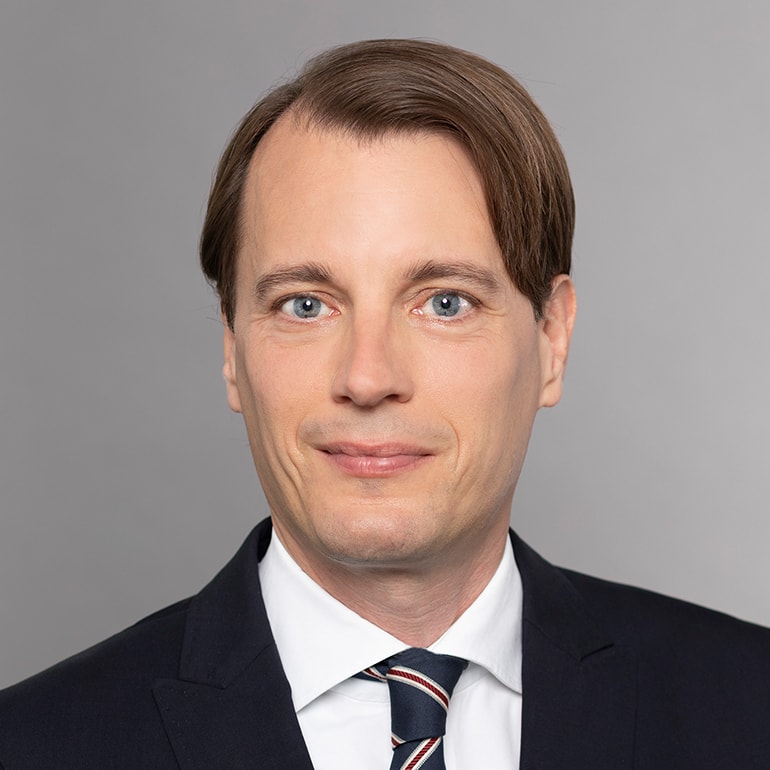 Das Bild zeigt einen Mann mit mittellangen, glatten Haaren und blauen Augen. Er trägt einen schwarzen Anzug mit einem weißen Hemd und einer gestreiften Krawatte. Der Hintergrund ist grau. Der Mann lächelt leicht und wirkt freundlich.