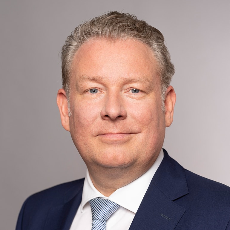 Ein höflich lächelnder Mann mit kurzen, blonden Haaren und einem hellblauen Anzug steht vor einem grauen Hintergrund. Er trägt ein weißes Hemd und eine hellblaue Krawatte. Der Ausdruck vermittelt Freundlichkeit und Professionalität.