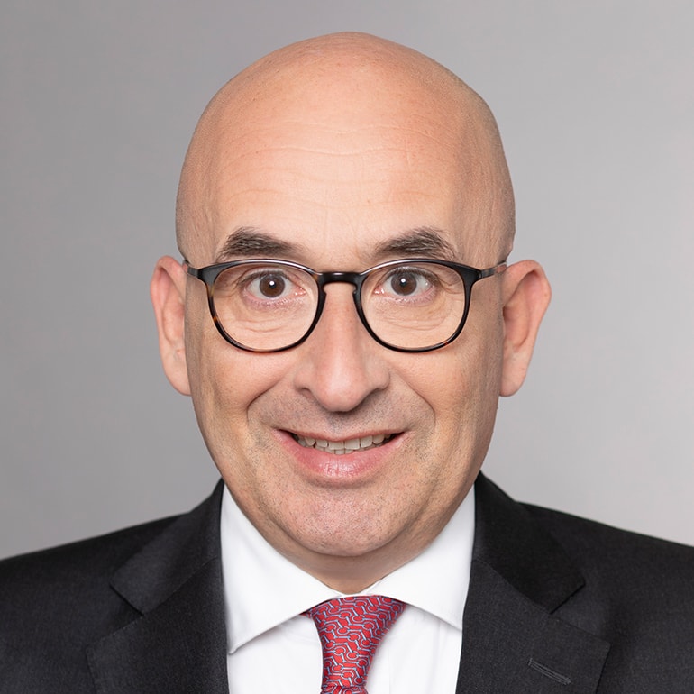 Ein mittelalterer Mann mit Glatze, der eine schwarze Anzugjacke und ein weißes Hemd mit roter, gemusterter Krawatte trägt. Er hat eine freundliche Ausstrahlung und lächelt direkt in die Kamera. Der Hintergrund ist neutral und hellgrau.