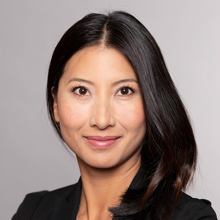 Ein Porträt einer Frau mit langen, dunklen Haaren. Sie trägt einen schwarzen Blazer und hat einen freundlichen Ausdruck auf ihrem Gesicht. Der Hintergrund ist einfarbig und hell, was die Frau in den Fokus rückt.