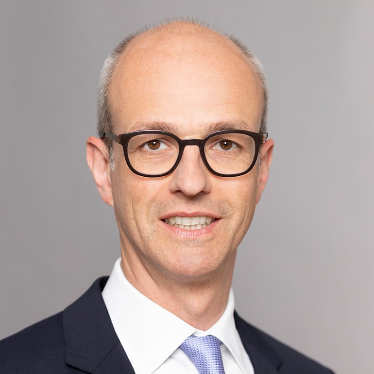 Porträt eines Mannes mit Glatze, der eine Brille trägt. Er lächelt freundlich und trägt einen dunklen Anzug mit einer hellen Krawatte und einem weißen Hemd. Der Hintergrund ist neutral grau.