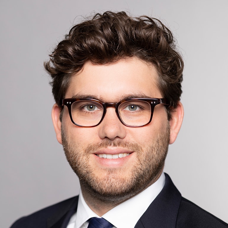 Ein junger Mann mit lockigem, braunem Haar und einem gepflegten Bart lächelt in die Kamera. Er trägt eine dunkle Anzugjacke und ein weißes Hemd mit einer blauen Krawatte. Die Hintergrundfarbe ist hell und neutral.