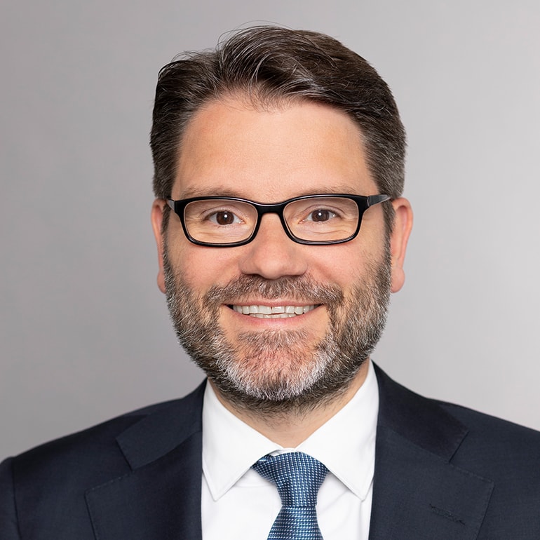 Porträt eines Mannes mit Brille und kurzem, braunem Haar. Er trägt einen Anzug mit Krawatte und lächelt freundlich. Der Hintergrund ist einfarbig und neutral.