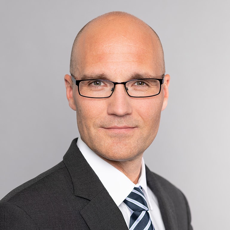 Ein Mann mit Glatze trägt eine schwarze Anzugjacke und ein gestreiftes Hemd mit Krawatte. Er blickt direkt in die Kamera und hat eine selbstbewusste Miene. Der Hintergrund ist neutral und hell.