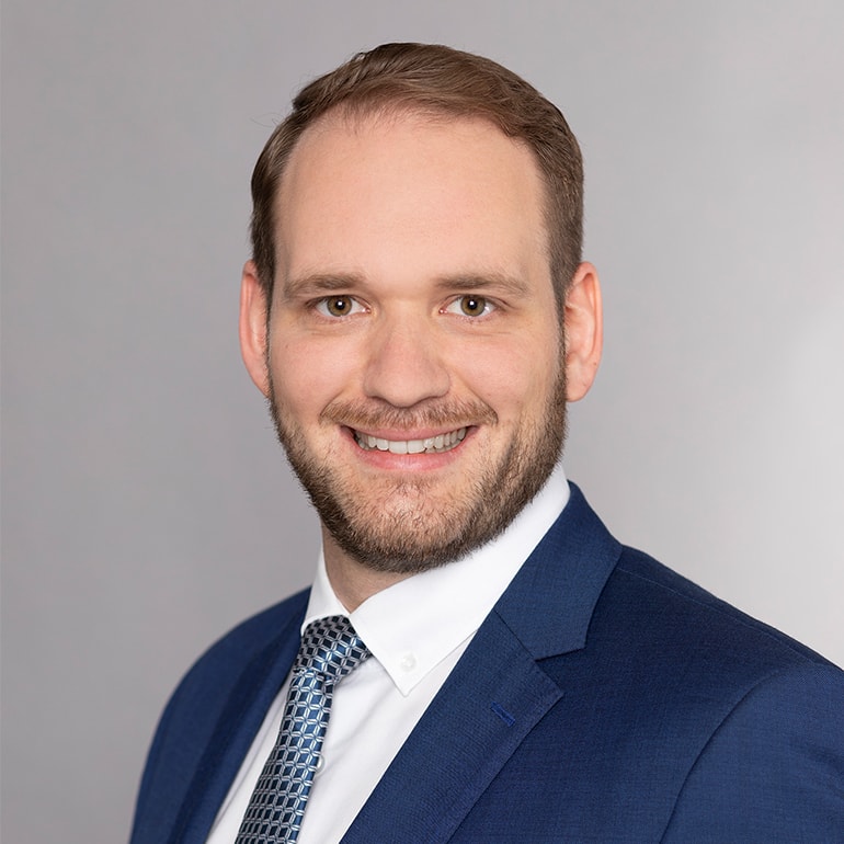 Ein lächelnder Mann mit einem kurzen Bart trägt einen blauen Anzug und eine passende Krawatte. Der Hintergrund ist grau und unifarben. Der Mann schaut direkt in die Kamera und vermittelt einen professionellen Eindruck.