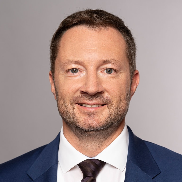 Porträt eines Mannes mit kurzen, braunen Haaren und einem gepflegten Bart. Er trägt einen dunkelblauen Anzug mit einem weißen Hemd und einer melierten Krawatte. Der Mann lächelt leicht und blickt direkt in die Kamera vor einem einfarbigen Hintergrund.