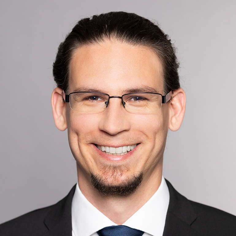 Porträt eines lächelnden Mannes mit dunklen, langen Haaren, die zu einem Zopf gebunden sind. Er trägt eine Brille, einen schwarzen Anzug und eine blaue Krawatte. Der Hintergrund ist einfarbig hellgrau. Sein Ausdruck vermittelt Freundlichkeit und Professionalität.