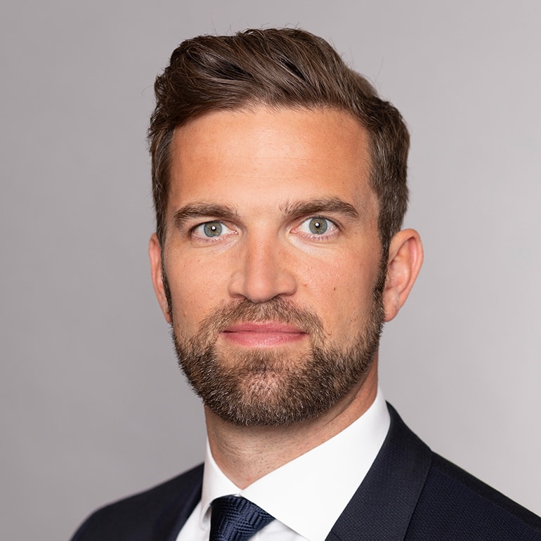 Porträt eines Mannes mit kurzem, braunem Haar und Vollbart. Er trägt einen dunkelblauen Anzug und ein helles Hemd. Der Hintergrund ist neutral grau. Der Mann schaut direkt in die Kamera und hat einen neutralen, aber selbstbewussten Gesichtsausdruck.