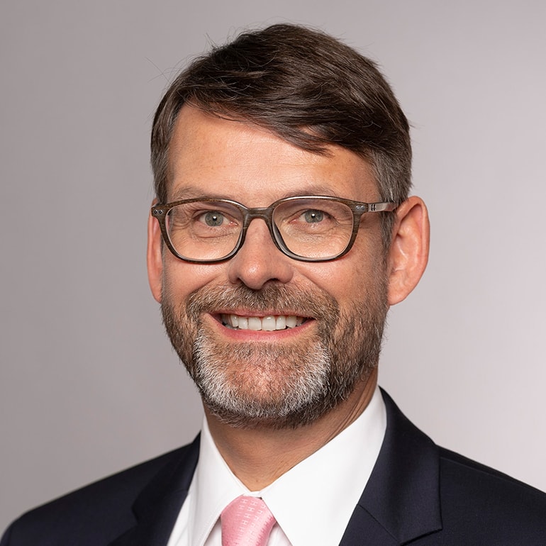 Porträt eines Mannes mit Brille und einem freundlichen Lächeln. Er trägt einen dunkelblauen Anzug und ein hellrosa Hemd. Der Hintergrund ist neutral und grau. Die Aufnahme vermittelt einen professionellen und einladenden Eindruck.