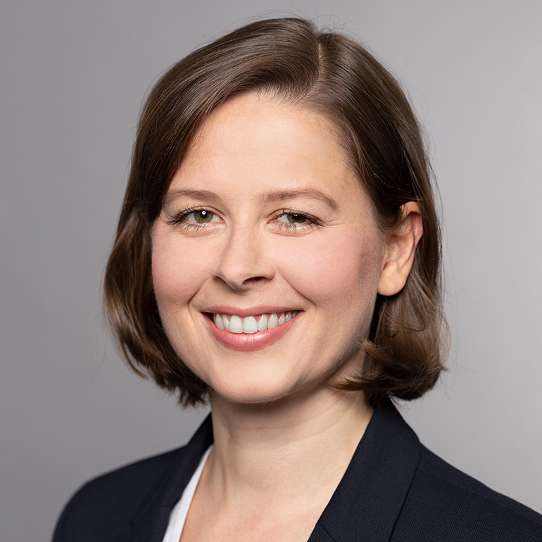 Porträt einer lächelnden Frau mit kurzen braunen Haaren, die einen dunklen Blazer trägt. Der Hintergrund ist neutral und grau, was den Fokus auf ihr Gesicht lenkt. Sie strahlt Freundlichkeit und Selbstbewusstsein aus.