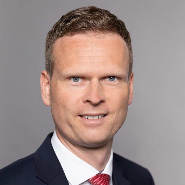 Porträt eines Mannes mit kurzem, hellbraunem Haar und blauem Anzug. Er trägt ein weißes Hemd und eine rote Krawatte. Der Hintergrund ist neutral und grau. Der Mann lächelt freundlich in die Kamera.