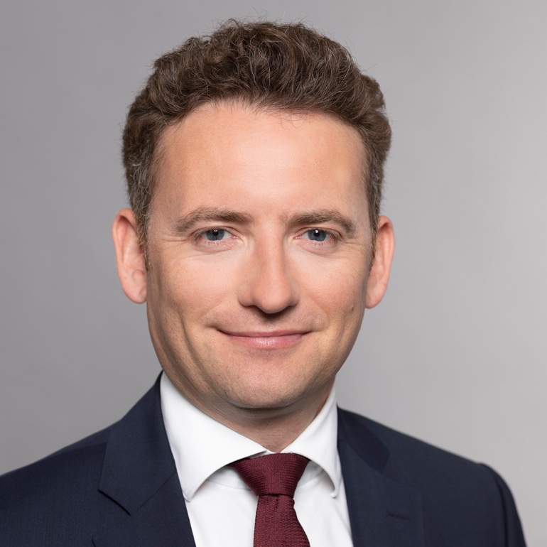 Ein Mann mit lockigem, dunkelbraunem Haar trägt einen dunklen Anzug und eine hellblaue Krawatte. Er lächelt freundlich in die Kamera vor einem grauen Hintergrund. Seine Augen sind blau und strahlen Vertrauen aus.