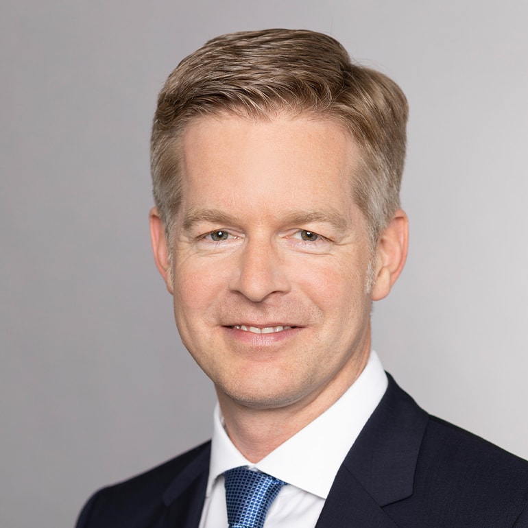 Ein lächelnder Mann mit blondem, kurz geschnittenem Haar trägt einen dunklen Anzug und ein hellblaues Hemd mit einer passenden Krawatte. Der Hintergrund ist hellgrau, was die Hauptmerkmale des Mannes betont.