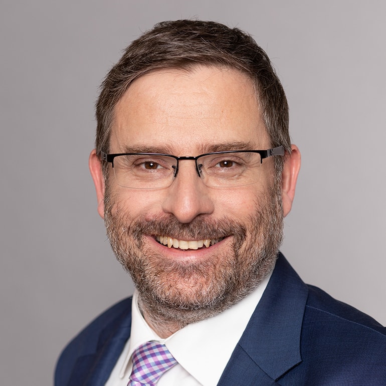 Porträt eines Mannes mit Brille und Bart, der freundlich lächelt. Er trägt einen dunklen Anzug mit einem karierten Krawatte. Der Hintergrund ist einfarbig grau. Das Bild vermittelt Professionalität und Freundlichkeit.