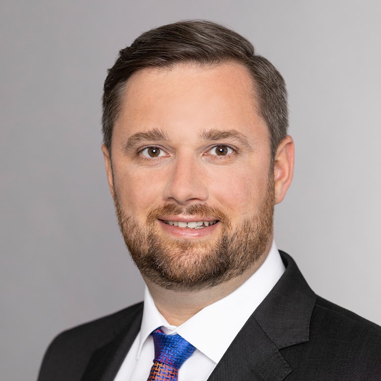 Ein lächelnder Mann mit kurzen, braunen Haaren und einem gepflegten Bart trägt einen schwarzen Anzug und eine bunte Krawatte. Der Hintergrund ist grau und unauffällig, um den Fokus auf sein Gesicht zu lenken.