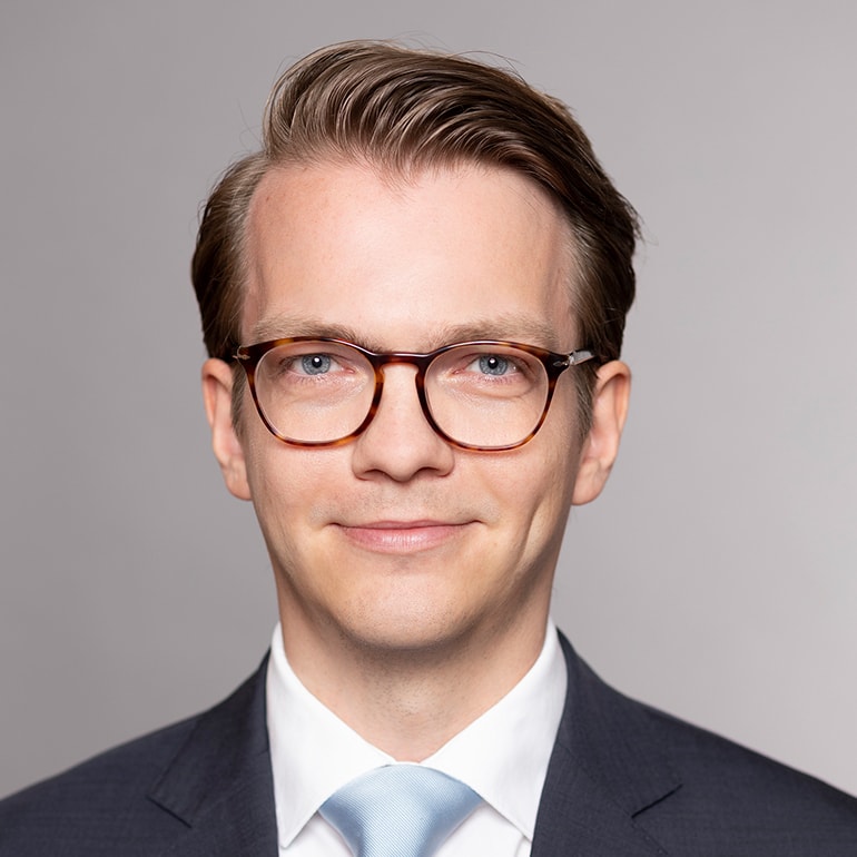 Porträt eines Mannes mit blonden Haaren, der eine Brille trägt. Er ist in einen dunklen Anzug mit hellblauer Krawatte gekleidet und lächelt freundlich. Der Hintergrund ist neutral und grau.