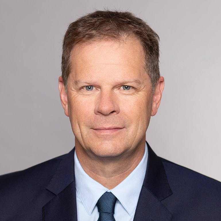 Porträt eines Mannes mit kurzen, hellen Haaren und blauem Anzug. Er hat ein leichtes Lächeln, trägt ein helles Hemd und eine Krawatte. Der Hintergrund ist einfarbig hellgrau. Die Ausstrahlung wirkt freundlich und professionell.