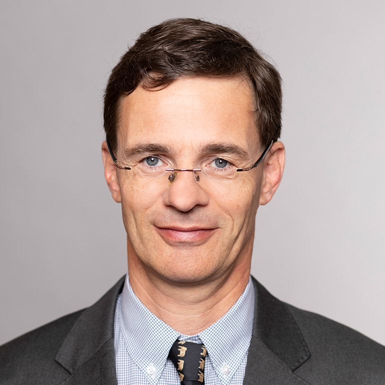 Porträt eines Mannes mit lässigem, freundlichem Ausdruck. Er trägt eine Brille, einen Anzug und eine Krawatte mit einem Tiermotiv. Der Hintergrund ist einfarbig und neutral.