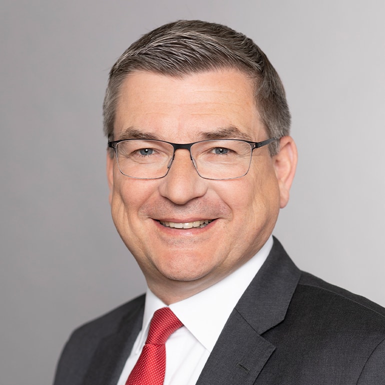 Porträt eines lächelnden Mannes mit Brille, kurzen grauen Haaren und einem Anzug. Er trägt eine rote Krawatte und hat einen freundlichen Gesichtsausdruck. Der Hintergrund ist neutral und hell.