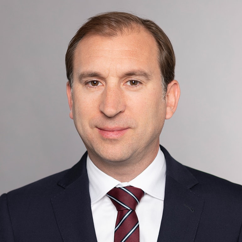 Porträt eines Mannes mit kurzem, braunem Haar, der einen Anzug mit Krawatte trägt. Er schaut direkt in die Kamera und hat einen neutralen Gesichtsausdruck. Der Hintergrund ist grau und einfarbig.