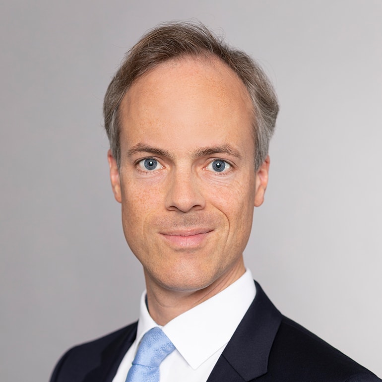 Ein lächelnder Mann mit blonden Haaren und heller Haut trägt ein dunkelblaues Jacket und eine hellblaue Krawatte. Der Hintergrund ist einfarbig grau. Seine Gesichtsausdrücke strahlen Freundlichkeit und Professionalität aus.