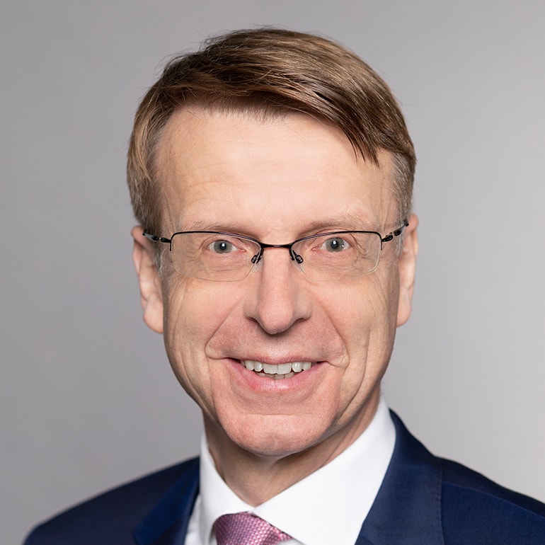 Porträt eines lächelnden Mannes mit Brille und kurzen braunen Haaren. Er trägt einen blauen Anzug und eine hellrosa Krawatte. Der Hintergrund ist grau und neutral gehalten.