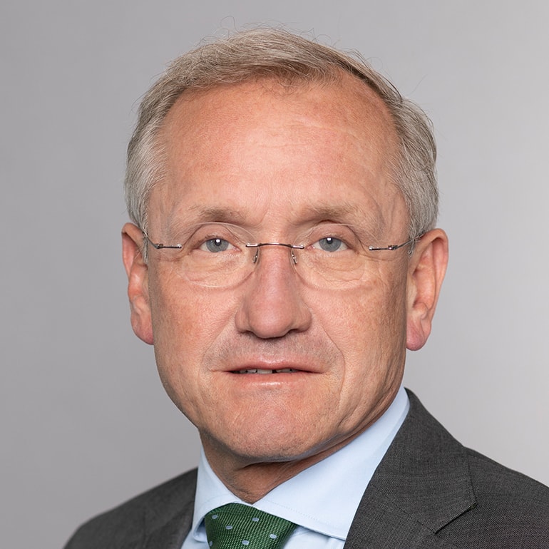 Porträt eines Mannes mit kurzen, grauen Haaren und Brille. Er trägt einen grauen Anzug mit einem blauen Hemd und einer grünen Krawatte mit Punkten. Der Hintergrund ist neutral und hellgrau. Der Mann blickt direkt in die Kamera.