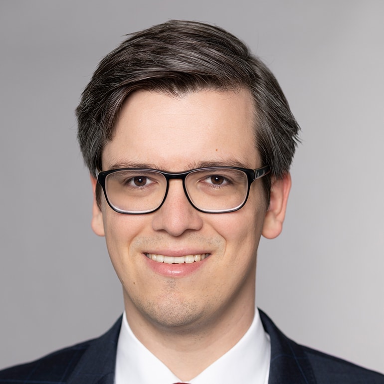 Ein junger Mann mit Brille und einem freundlichen Lächeln. Er trägt einen dunklen Anzug mit einer hellen Krawatte und hat kurzes, braunes Haar. Der Hintergrund ist neutral gehalten, was den Fokus auf sein Gesicht lenkt.