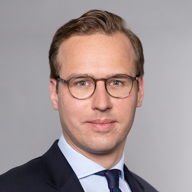 Eine nahaufgenommene Porträtaufnahme eines Mannes mit Brille. Er trägt einen dunklen Anzug und ein hellblaues Hemd mit einer gepunkteten Krawatte. Der Hintergrund ist grau und lenkt nicht von seinem Gesicht ab, das einen neutralen Ausdruck zeigt.