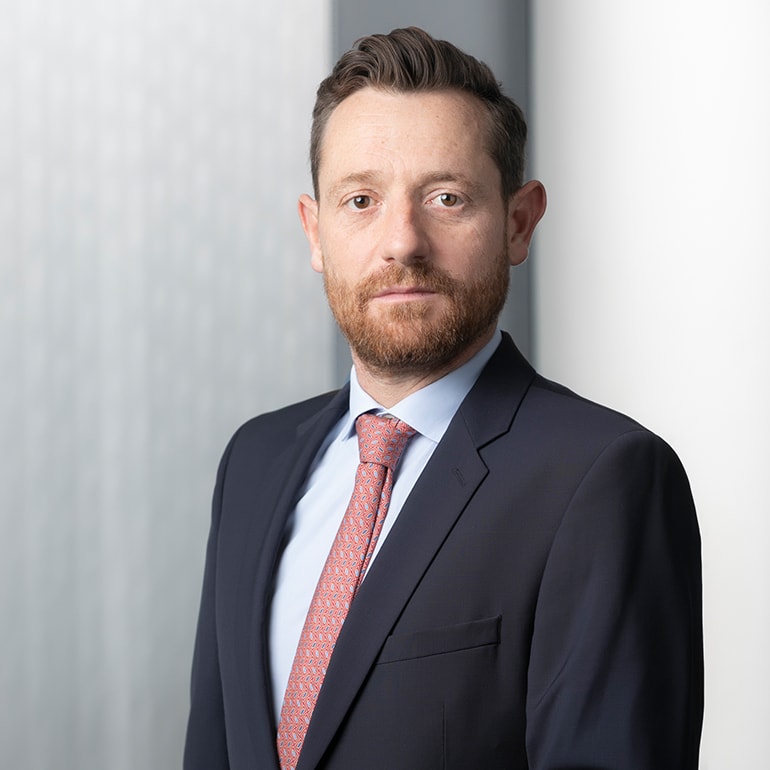 Ein mittelalterlicher Mann mit einem Bart trägt einen dunklen Anzug und ein hellblaues Hemd, ergänzt durch eine gemusterte Krawatte. Er steht vor einem unscharfen, hellen Hintergrund und blickt direkt in die Kamera, was Selbstbewusstsein und Professionalität vermittelt.