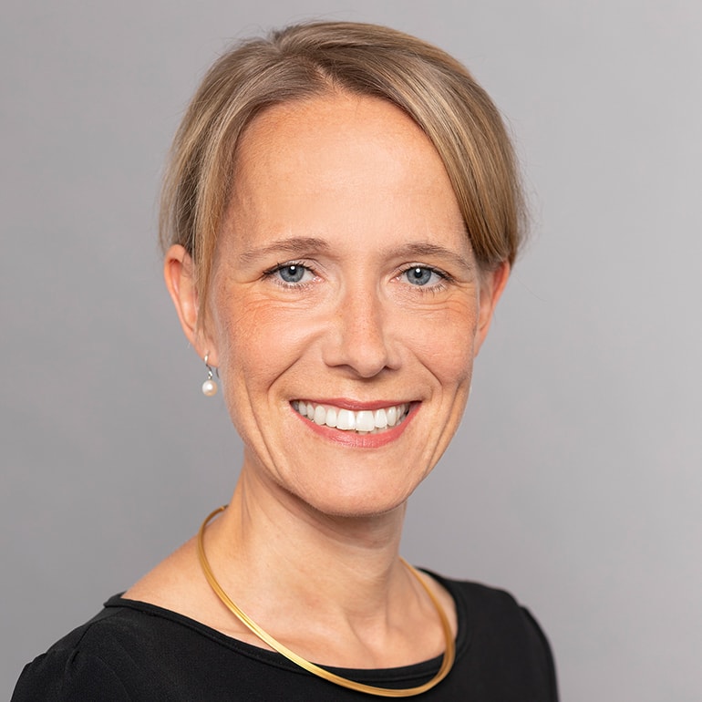 Ein Porträt einer lächelnden Frau mit blonden, schulterlangen Haaren. Sie trägt ein schwarzes Oberteil und eine goldene Halskette. Ihr Gesicht zeigt freundliche Züge, und sie hat blaue Augen. Der Hintergrund ist grau.