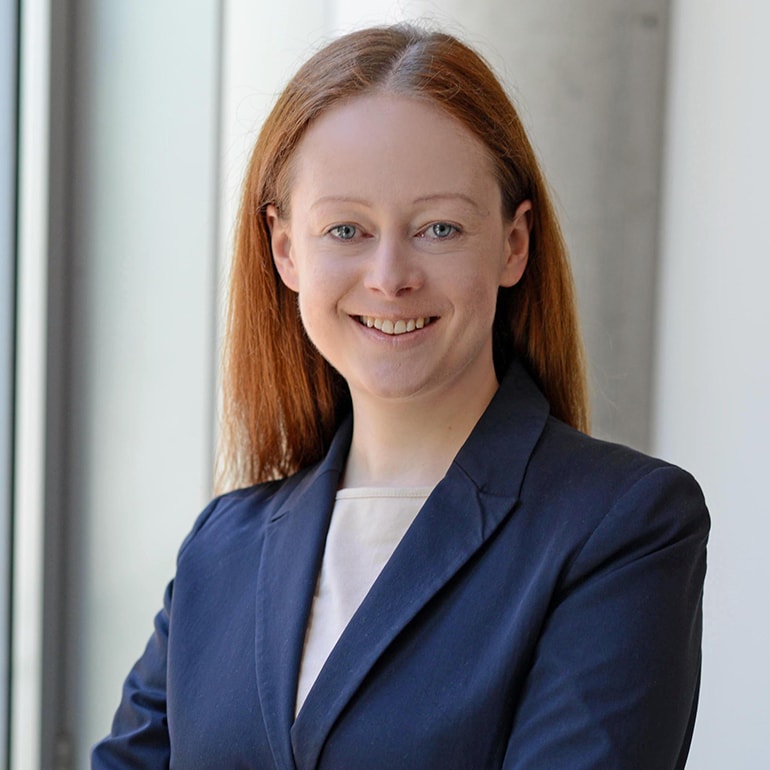 Eine lächelnde Frau mit langen, rotbraunen Haaren trägt einen dunklen Blazer und ein helles Oberteil. Sie steht vor einem Fenster mit natürlichem Licht. Ihr Gesichtsausdruck strahlt Freundlichkeit und Selbstbewusstsein aus.