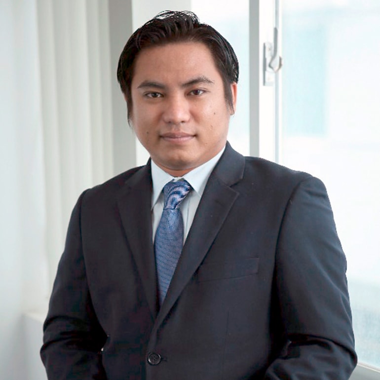 Ein Mann steht in einem Anzug mit Krawatte und leichtem Lächeln vor einem Fenster. Er hat kurze, dunkle Haare und eine ernste Miene. Der Hintergrund ist hell und minimalistisch, was den Fokus auf die Person lenkt.